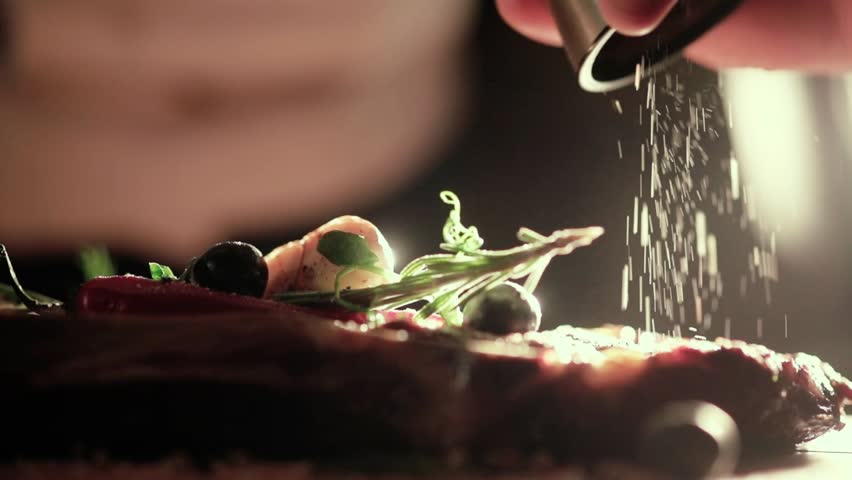 Extreme Close Up view of a Hand Seasoning Cooked piece of Meat in a Bright Light. Chef Accompanying Steak with Garlic, Rosemary, Pepper, Salt. Delicious, Modern Cuisine, Exclusive Recipe. 120 fps.